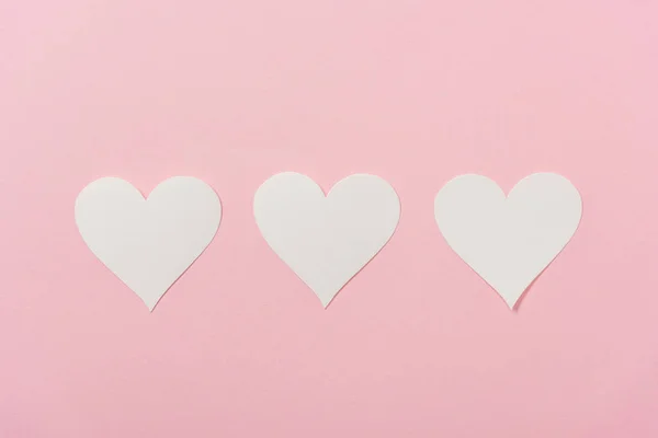 Corazones de papel blanco dispuestos en fila sobre fondo rosa - foto de stock