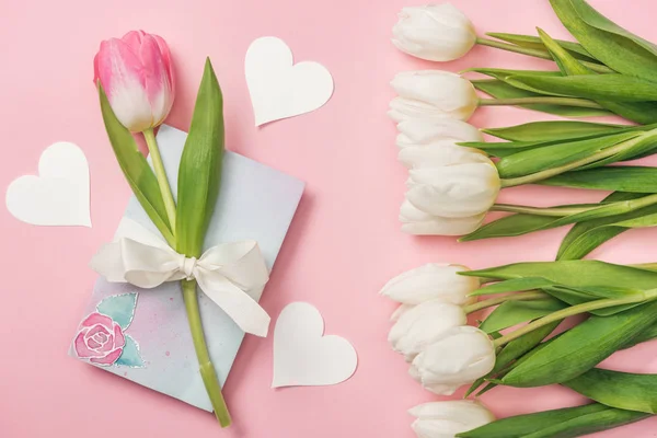 Tulipano rosa con fiocco bianco su biglietto di auguri, cuori di carta e tulipani bianchi su sfondo rosa — Foto stock