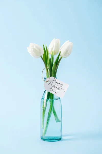 Buquê de tulipas brancas em vaso de vidro com mães felizes etiqueta dia saudação no fundo azul — Fotografia de Stock