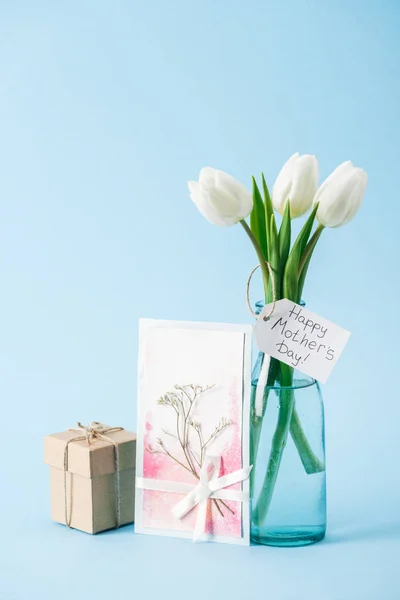 Happy mothers day greeting card, bouquet of white tulips with paper label, and gift box on blue background — Stock Photo