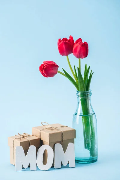 Boîtes-cadeaux, bouquet de tulipes rouges et mot de papier blanc maman sur fond bleu — Photo de stock