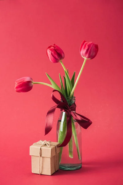 Caja de regalo y ramo de tulipanes rojos con cuenco de satén rojo en jarrón de vidrio sobre fondo rojo - foto de stock