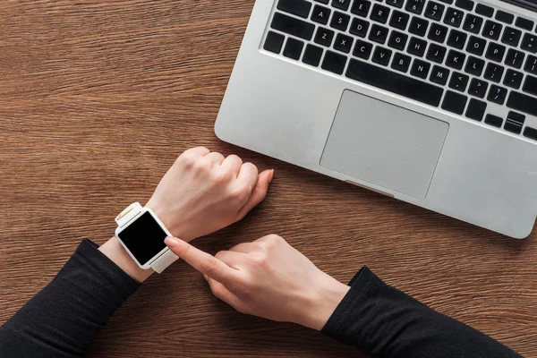 Vista recortada de la mujer con el ordenador portátil y smartwatch con pantalla en blanco posando sobre fondo de madera - foto de stock