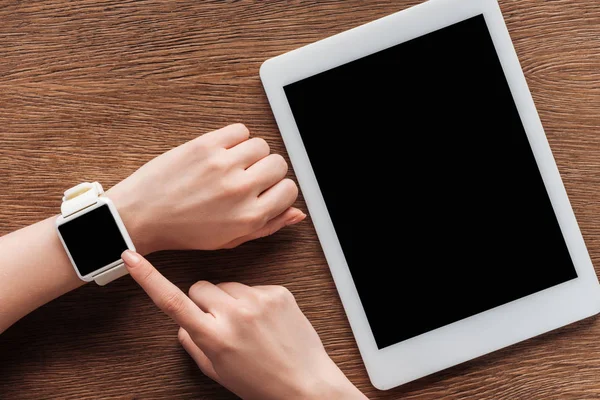 Vista parcial de mujer con tablet digital y smartwatch con pantalla en blanco sobre fondo de madera - foto de stock