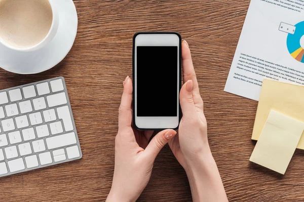 Vista parcial de la mujer sosteniendo el teléfono inteligente con pantalla en blanco en el lugar de trabajo - foto de stock