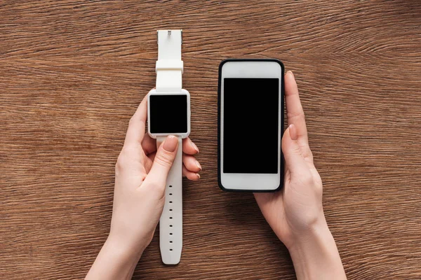 Partial view of woman holding smartwatch and smartphone with blank screen on wooden background — Stock Photo
