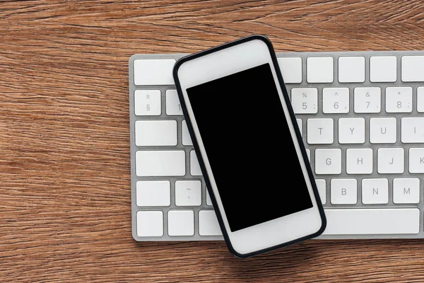 Vista superior del teclado y smartphone con pantalla en blanco sobre fondo de madera - foto de stock