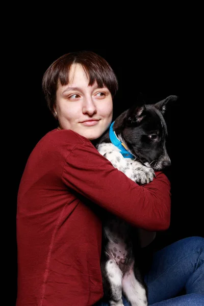 Mujer en suéter rojo abrazando y sosteniendo perro mestizo en collar azul aislado en negro — Stock Photo