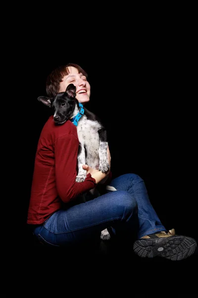 Femme en pull rouge et jeans assis, étreignant et tenant chien bâtard en col bleu isolé sur noir — Photo de stock