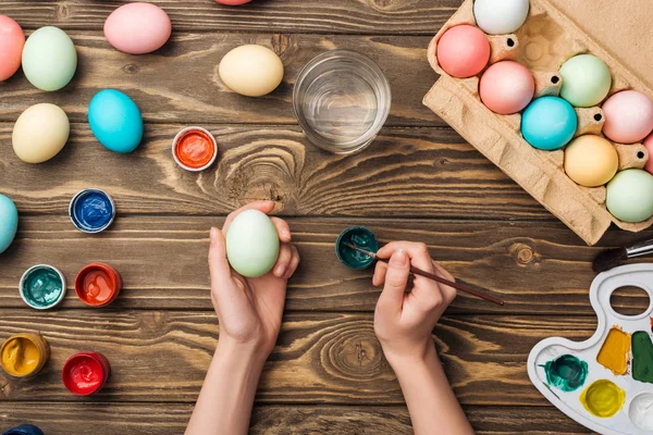 Vista parziale di ragazza decorazione uova di Pasqua con pennello a tavolo in legno con vernici e tavolozza dei colori — Foto stock