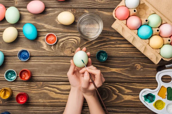 Vista parziale di ragazza decorazione uova di Pasqua con pennello a tavolo in legno con vernici e tavolozza dei colori — Foto stock
