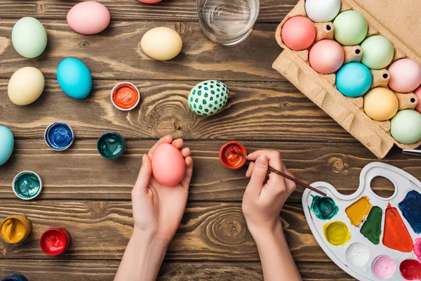 Частковий вид дівчини, що прикрашає великодні яйця за дерев'яним столом з фарбами та кольоровою палітрою — стокове фото