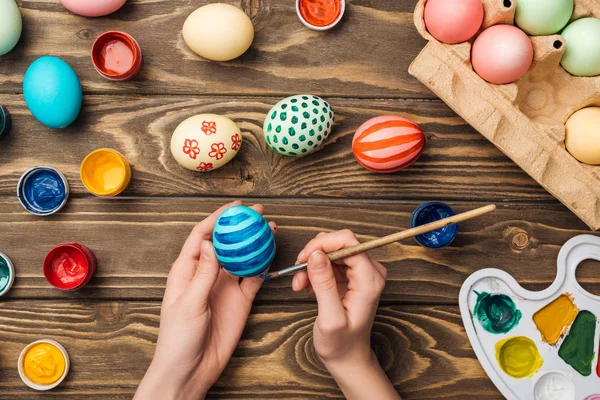 Верхний вид женщины, рисующей пасхальные яйца за деревянным столом с красками и цветовой палитрой — стоковое фото