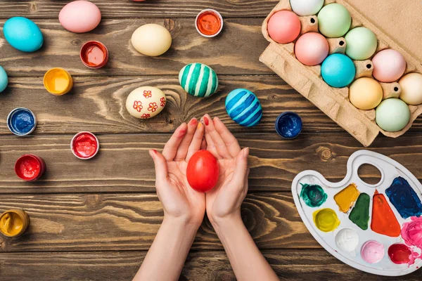 Vue du dessus de la femme tenant un oeuf de Pâques peint à une table en bois avec des peintures et une palette de couleurs — Photo de stock