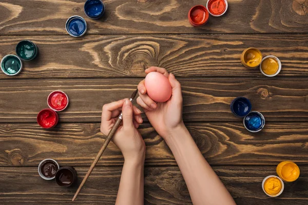 Верхний вид женщины, рисующей пасхальное яйцо за деревянным столом красками — стоковое фото