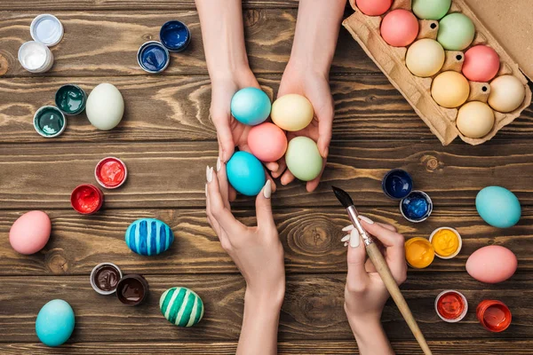Vista superior de las mujeres sosteniendo y pintando huevos de Pascua en la mesa de madera - foto de stock