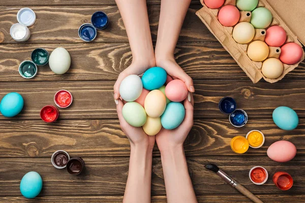 Top view of women holding pastel easter eggs at wooden table with paints — Stock Photo