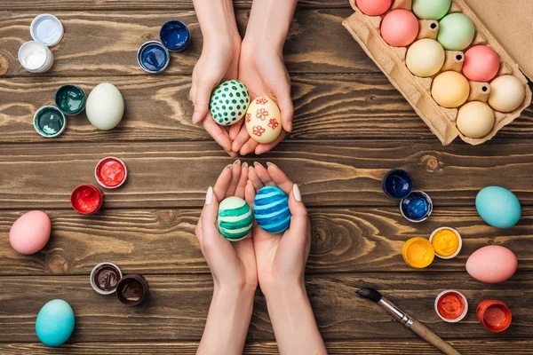 Top view of women holding ornamental easter eggs at wooden table with paints — Stock Photo