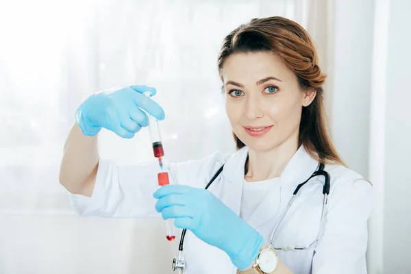 Médico sorridente em luvas de látex segurando seringa com amostra de sangue e tubo de ensaio — Fotografia de Stock