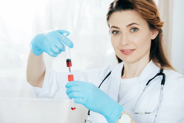 Sonriente doctor en guantes de látex sosteniendo jeringa con muestra de sangre y probeta - foto de stock