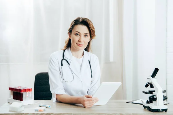 Médecin souriant en manteau blanc avec stéthoscope en utilisant une tablette numérique en clinique — Photo de stock