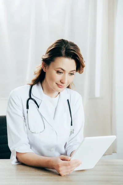Médico sonriente de bata blanca con estetoscopio usando tableta digital en la clínica - foto de stock