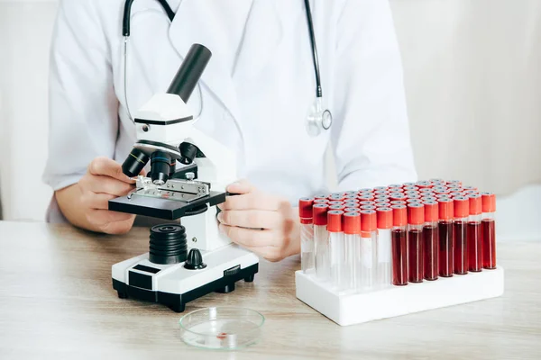 Vista recortada del médico de bata blanca con estetoscopio utilizando microscopio - foto de stock