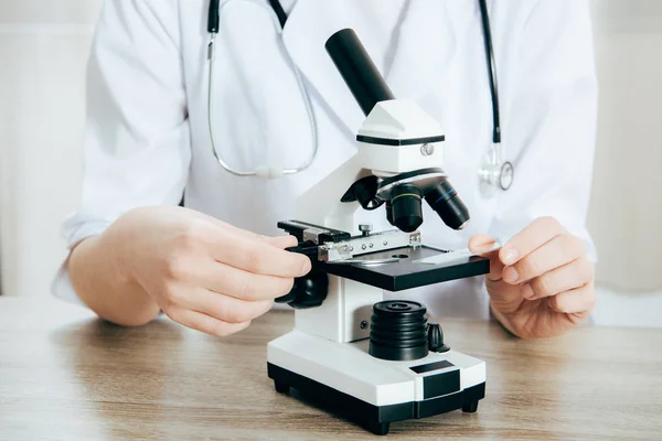 Cropped view of doctor in white coat with stethoscope using microscope — Stock Photo
