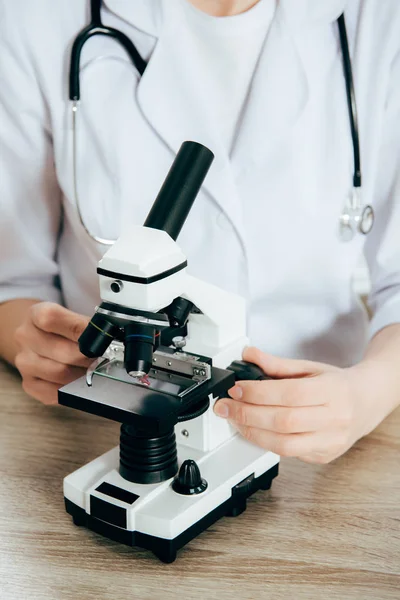 Vista recortada del médico de bata blanca con estetoscopio utilizando microscopio - foto de stock