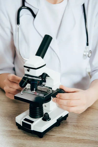 Cropped view of doctor in white coat with stethoscope using microscope — Stock Photo