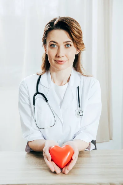 Médico sonriente de capa blanca con estetoscopio que sostiene el corazón de plástico - foto de stock