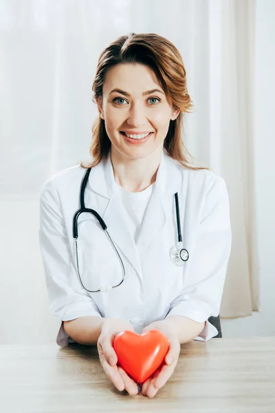Médico sorridente em casaco branco com estetoscópio segurando coração de plástico — Fotografia de Stock