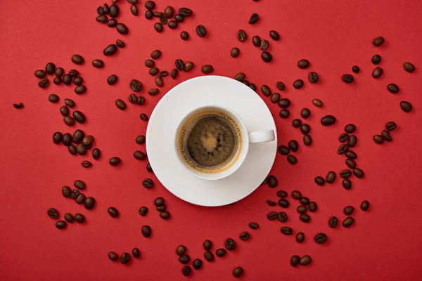 Вид зверху смачної кави в чашці біля розкиданих обсмажених бобів на червоному тлі — стокове фото