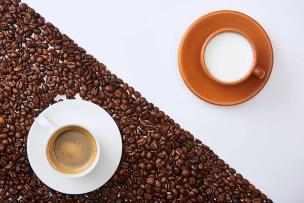 Vue du dessus de délicieux café dans la tasse parmi les grains torréfiés près de tasse de lait sur fond blanc — Photo de stock