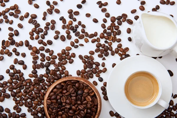 Blick von oben auf Kaffee in Tasse auf Untertasse in der Nähe verstreuter gerösteter Bohnen, Holzschale und Milchkanne — Stockfoto