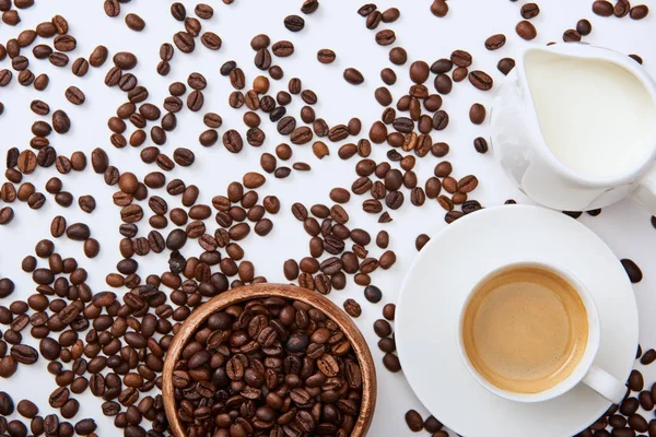 Top view of coffee in cup on saucer near scattered roasted beans, wooden bowl and milk jug — Stock Photo