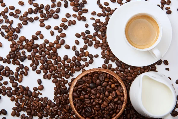 Top view of tasty coffee in cup on saucer near scattered roasted beans, wooden bowl and milk jug — Stock Photo