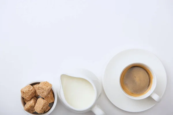 Вид зверху смачної кави в чашці на блюдці біля молока і коричневого цукру на білому тлі — стокове фото