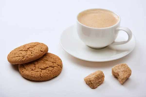 Leckeren Kaffee mit Schaum in Tasse auf Untertasse in der Nähe von braunem Zucker und Kekse auf weißem Hintergrund — Stockfoto