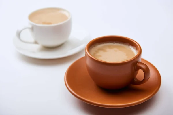 Messa a fuoco selettiva di caffè con schiuma in tazze su piattini su sfondo bianco — Foto stock