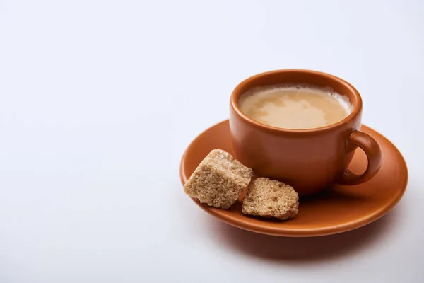 Café saboroso com espuma na xícara no pires perto de açúcar mascavo no fundo branco com espaço de cópia — Fotografia de Stock