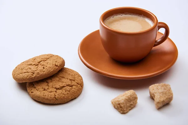 Café saboroso com espuma na xícara no pires perto de açúcar mascavo e biscoitos doces no fundo branco — Fotografia de Stock