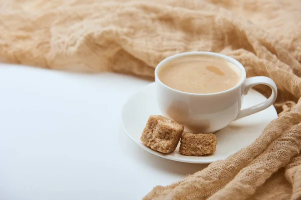Délicieux café avec mousse en tasse blanche sur soucoupe avec sucre brun près du tissu sur fond blanc — Photo de stock