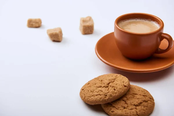 Köstlicher Kaffee mit Schaum in brauner Tasse auf Untertasse mit braunem Zucker und Keksen auf weißem Hintergrund — Stockfoto