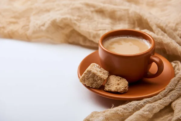 Délicieux café avec mousse dans une tasse sur soucoupe avec sucre brun près du tissu sur fond blanc — Photo de stock