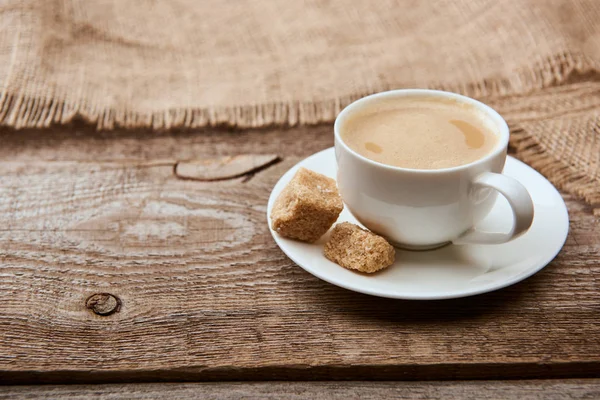 Delicioso café com espuma na xícara no pires com açúcar mascavo perto de pano de saco no fundo de madeira — Fotografia de Stock