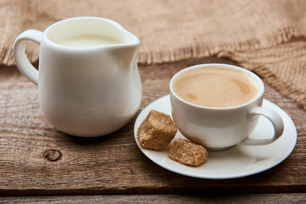 Delicioso café com espuma na xícara no pires com açúcar mascavo perto de pano de saco e jarro de leite no fundo de madeira — Fotografia de Stock