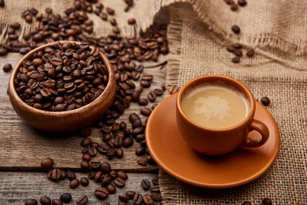 Chicchi di caffè in ciotola vicino a tazza di caffè su sfondo di legno — Foto stock