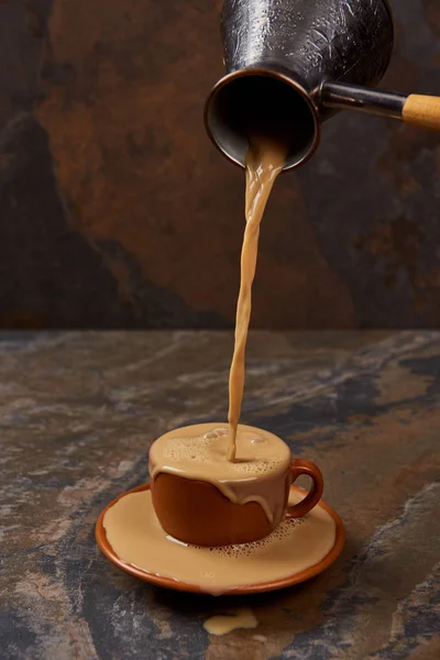 Verser le café chaud de cezve dans la tasse avec la flaque sur la soucoupe sur la surface de marbre — Photo de stock