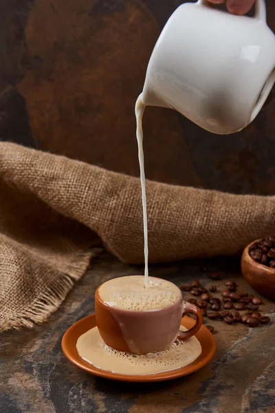 Vista cortada da pessoa derramando leite na xícara com café na mesa de mármore perto de pano de saco — Fotografia de Stock